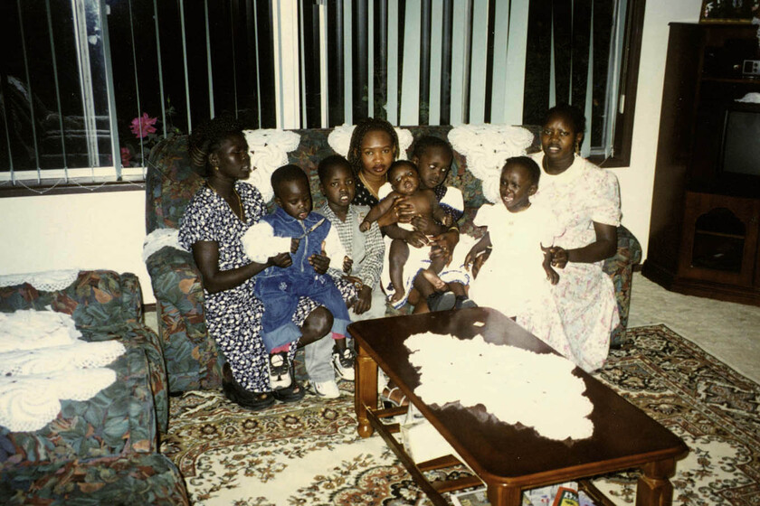 A living room with three young women in dresses seated on a couch, with four young children and one baby across their laps. There is a coffee table in front with a patterned rug on the floor, brightly coloured green and flower pattern couch, and Venetian blinds behind..