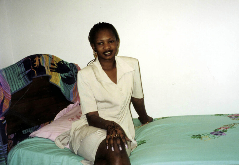 Young woman in a white suite top and skirt, sitting on a bed with a bright bright green bed spread, and brightly coloured towel hanging over a wooden bedhead.