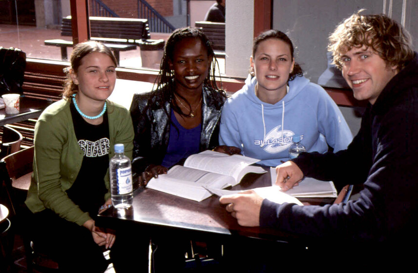 Three young women and one man seated smiling, at a small wooden table in a cafe, with books open on the table in front.