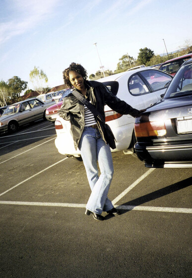 Young woman in jeans, striped top and leather jacket with satchel bag crossed over the body, standing in a car park leaning on the boot or trunk of a black car. There is a white car behind.