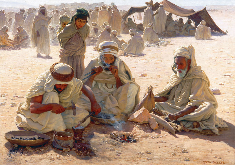 Group of three men seated in sand around a fire, wearing Arab-style clothing and head coverings. A young boy stands behind. In the background is a larger group of people, sitting in sunlight and under tents.