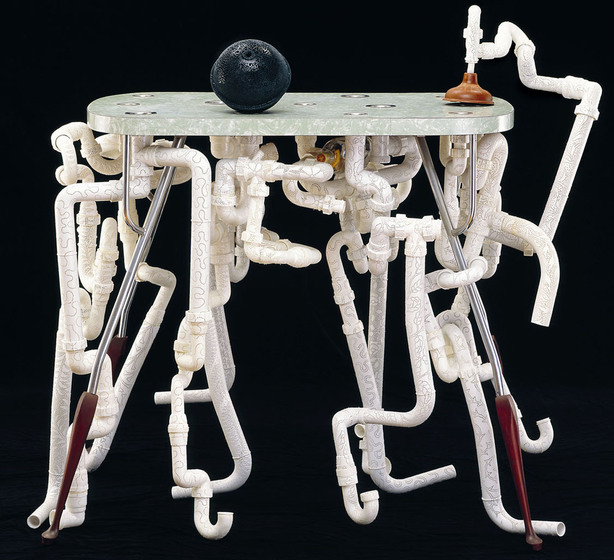 Small laminate kitchen table with a black ball and plunger on top, and a series of pipes attached beneath.