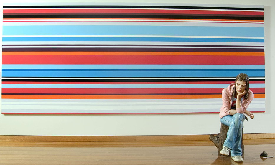 Two artworks, a large canvas with red, black, white and blue horizontal stripes features behind a standing sculpture of a woman seated on a log wearing blue jeans and a pink hooded jacket.