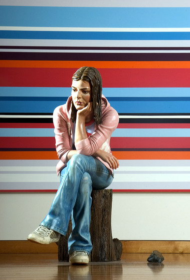 Standing sculpture of a woman seated on a log wearing blue jeans and a pink hooded jacket. Behind is a partial view of another artwork, a large canvas with red, black, white and blue horizontal stripes.