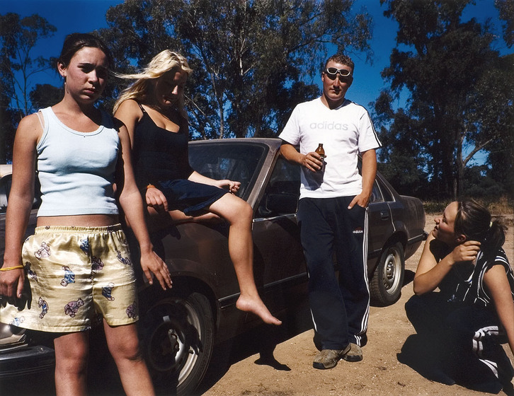 A brown car on sandy ground, with gum trees and blue sky seen behind. A woman in a black dress is seated on the hood or bonnet, with a teenage girl in shorts and a singlet to the left. A man leans on the car side, with a bottle of beer in his hand and sunglasses pushed up to his forehead. Another teenage girl is crouched to the right. 