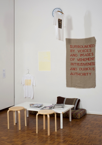 Corner of a white room with parquetry floor, with a clock on the wall and a tee-shirt handing in front. A second tee-shirt hangs lower down on the wall, with ayellow rectangle and hessian material featuring the text 'surrounded by voices and images of vehement intrusiveness and dubious authority'. In front on the floor is a low white table, with two wooden stools and a small brown mattress and cushion. There are papers on the table.