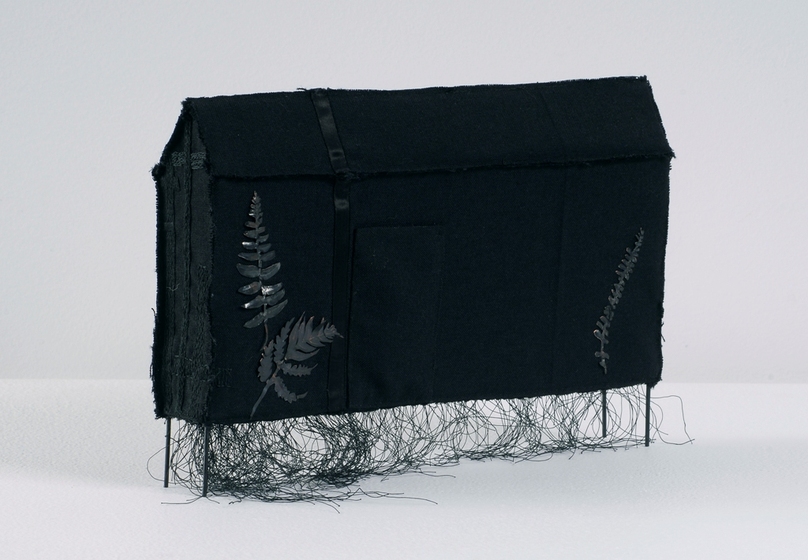 Wire covered with black fabric, shaped to form a long thin house shape on stilts. There are black painted fern leaves fixed to one side.