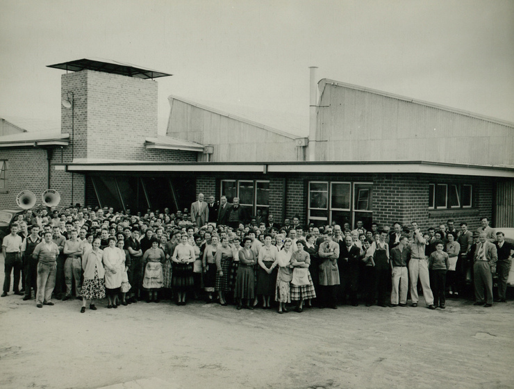 Black and white photograph of a large group of people, both men and women, standing in front of a brick building.