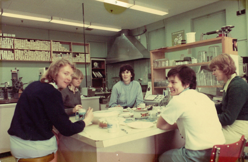 Colour photograph of five young women seated around a table in a factory or lab room. There are shelves behind featuring items of equipment.