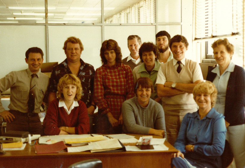 Colour photograph of a small group of men and women wearing 1970s clothing, seated and standing in an office behind a timber desk. 