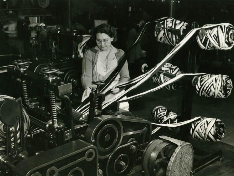 Woman seated behind at a large machine holding six large rolls of textile, being pulled through the machine's mechanics.