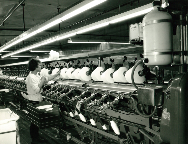 Large machine holding a row of large spools of white thread, being threaded from bars holding thread below. There is a woman standing in front of the machine, with arm held up to one of the spools. 