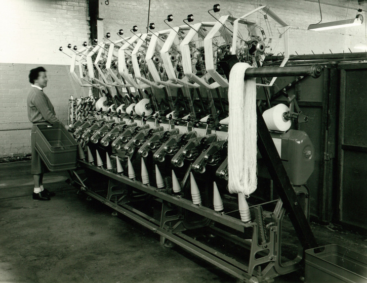 Large machine with repeating sections of thread being wound around in hexagon shapes, a woman with a large tub stands beside the machine.