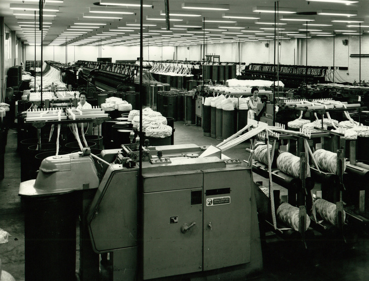Large factory room showing repeating  machines holding wound thread. There are two people standing amongst the machines.