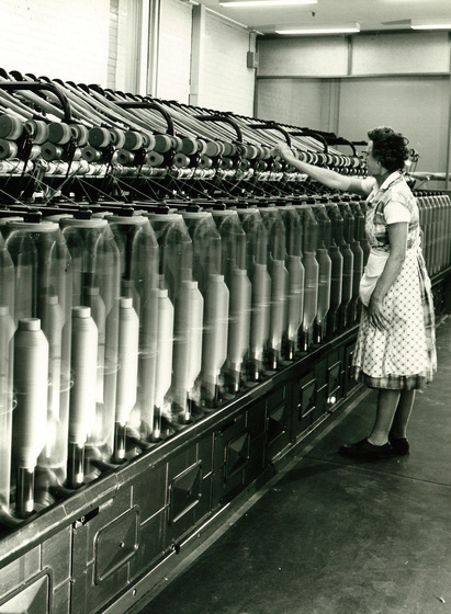 Middle aged woman in a polka dot apron standing in front of a long machine with repeating sections, with one arm raised up on the machinery.
