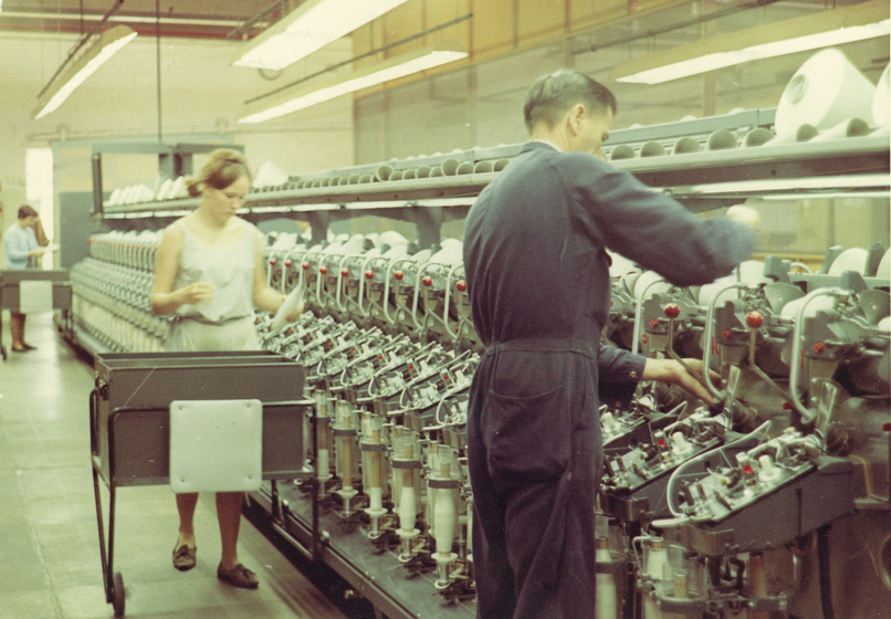 Man in a navy blue jumpsuit stands in front of a long machine with repeating sections. To his left is a young woman with a cart in front of her.