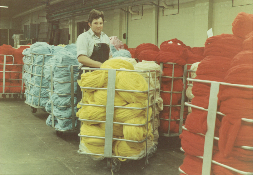 Man standing amongst metal frame open storage containers on wheels, each holding rolls of wound coloured thread in light blue, yellow and red.
