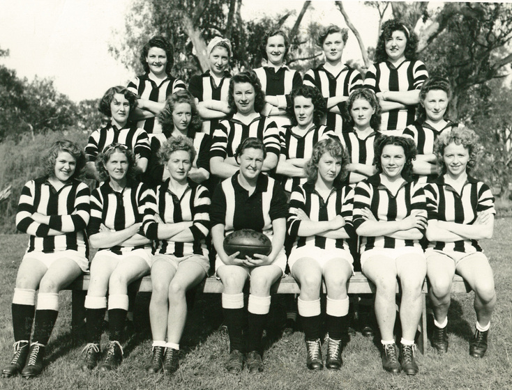 Group team photograph of three row of young women wearing black and white vertical striped Australian football guernseys. The woman seated front row in centre, holds an Australian football in her lap.