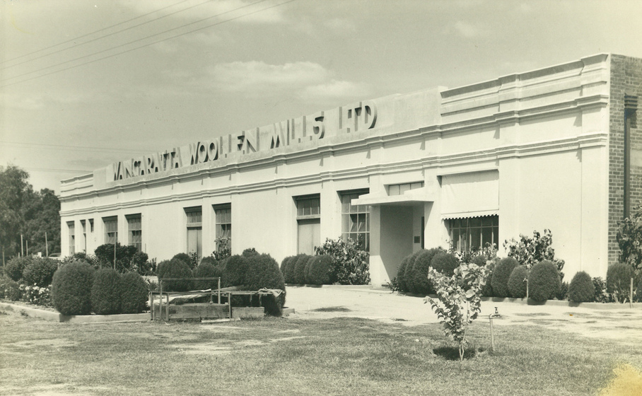 White building with rows of bushes in the garden in front, with the words 'Wangaratta Woollen Mills Ltd' at the top of the front of the building.