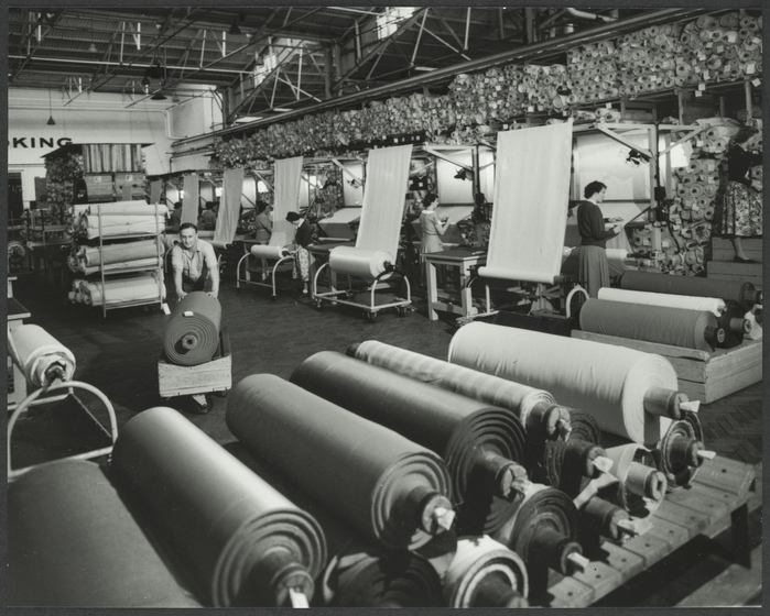 Interior of a factory room, featuring rolls of fabric both stacked in the foreground, and stored on racks and shelves behind. A man is centre, pushing a roll of fabric on a cart.