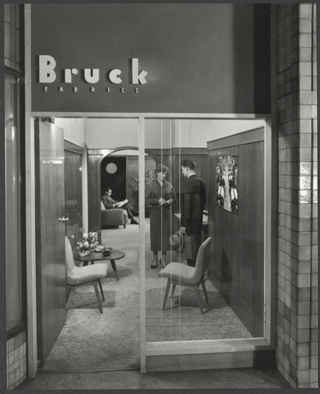 Shop front door and window, looking through into front room. This room is furnished like a living room, and above the front door is the text 'Bruck Fabrics'. There are a man and a woman standing inside, and another man is seated further in.