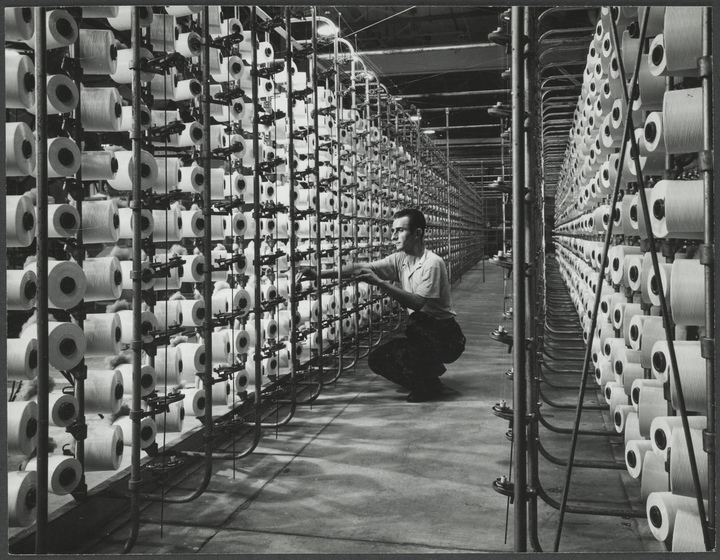 Man crouching in between two rows of stacked large spools of white thread.