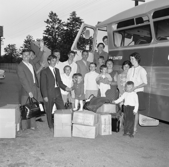 Group of men, women and children standing in front of a bus, with boxes and bags around their feet.