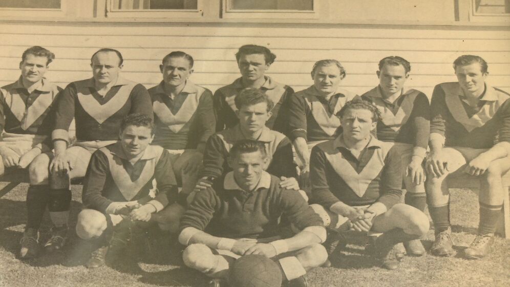 Group team photograph of two rows of men, plus a single man seated in front, wearing Australian football guernseys of dark colour with a light coloured chevron on the chest. The man seated front has a Australian football in front of him at his feet.
