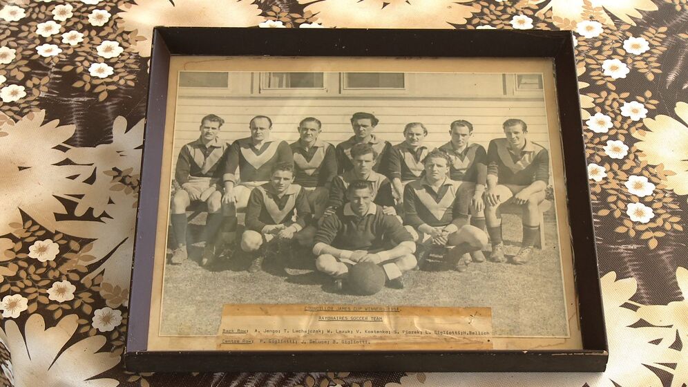 Framed team photograph sitting on a printed brown and cream floral surface, The photograph is Group team photograph of two rows of men, plus a single man seated in front, wearing Australian football guernseys of dark colour with a light coloured chevron on the chest. The man seated front has a Australian football in front of him at his feet.