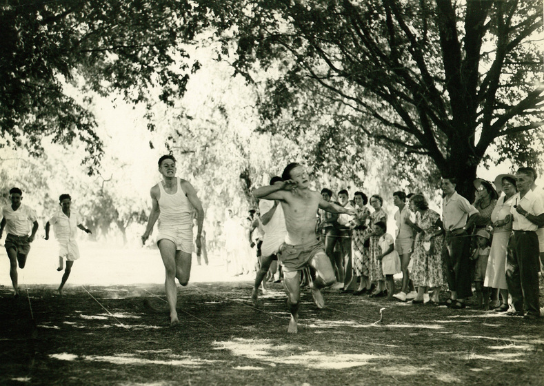 Five men are running barefoot across grass with lines marked in string, with trees behind and a crowd of people watching to the right.