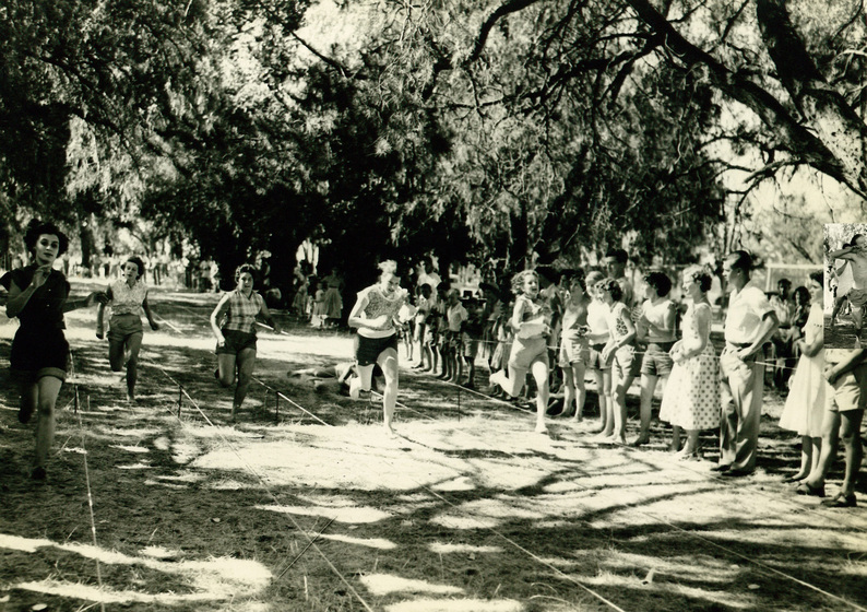 Five women are running barefoot across grass with lines marked in string, with trees behind and a crowd of people watching to the right.