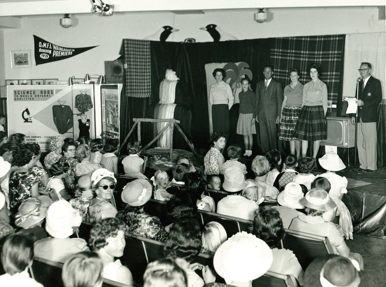 Seated audience in front of a small stage with six men and women standing in strong of a hung backdrop of fabric. 