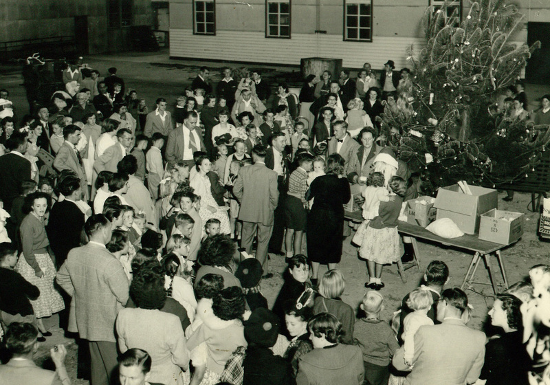 Large group of people standing outside around a table with boxes on top and with a decorated pine or Christmas tree behind.