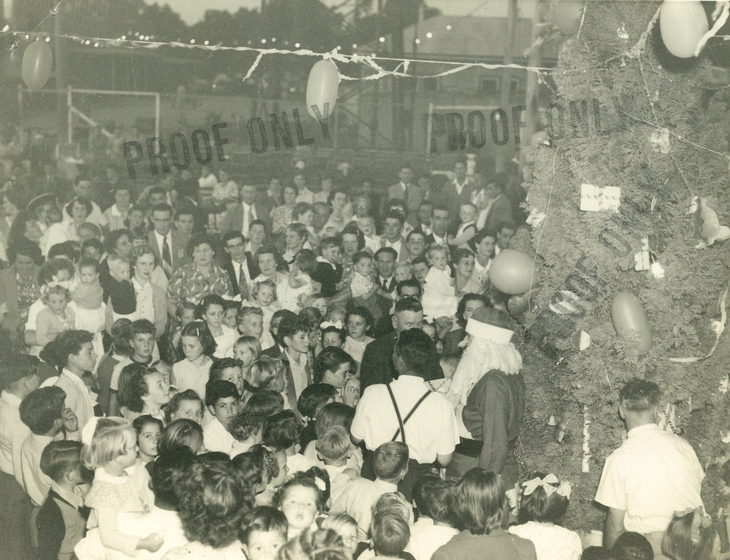 Large group of people, including children at the front, standing outside with a decorated pine or Christmas tree behind. Stamped on the image is the text 'proof only' three times.