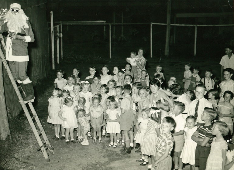 Group of children standing outside, with a man dressed as Santa Claus, or Father Christmas wearing sunglasses and standing up a ladder leaned on a fence.