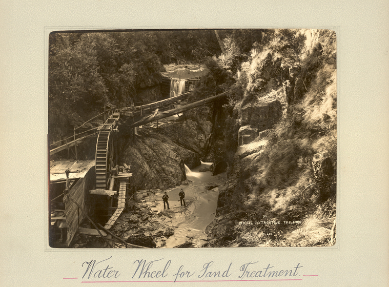 Black and white photograph of a gully with a waterfall centre, with timber structures built in front, including water wheel. Two men stand at the base of the gully. Text on the surrounding photo mount reads 'Water Wheel for Sand Treatment'. 