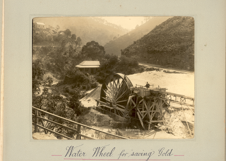 Black and white photograph of a wooden water wheel with a timber gantry sitting in a shallow gully between tree-covered hills. A house is seen behind, and a man stands on the gantry. Text on the surrounding photo mount reads 'Water Wheel for saving Gold'.