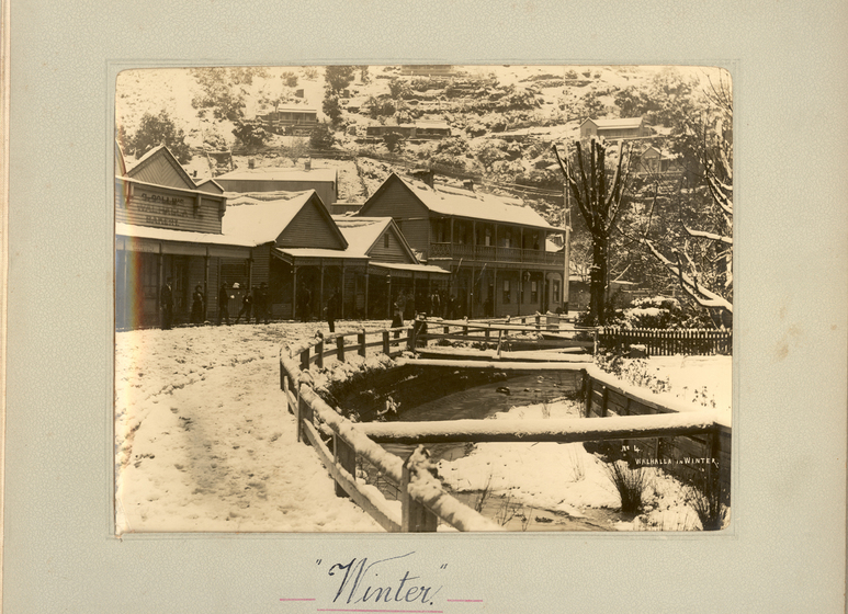 Black and white photograph of township along a frozen river, with timber buildings and the ground in front covered with snow.  Text on the surrounding photo mount reads 'Winter'. 