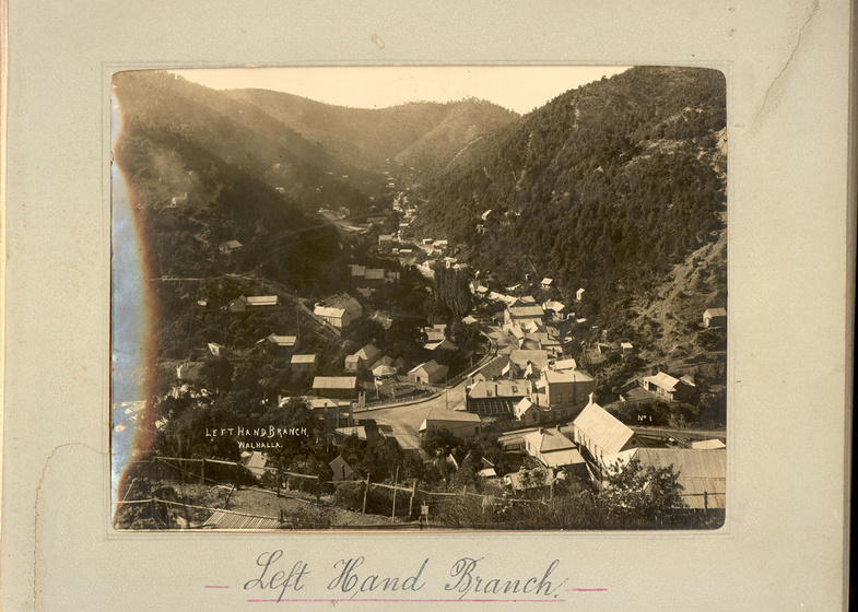 Black and white landscape photograph of a township set in a shallow gully, with tree-covered hills either side. There is a road going through the centre of town.  Text on the surrounding photo mount reads 'Left Hand Branch'