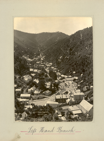 Black and white portrait photograph of a township set in a shallow gully, with tree-covered hills either side. There is a road going through the centre of town.  Text on the surrounding photo mount reads 'Left Hand Branch'