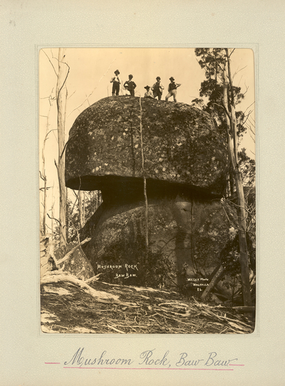 Black and white portrait photograph of two large boulders on top of each other, with five men standing on top of the top boulder. Text on the surrounding photo mount reads 'Mushroom Rock, Baw Baw'
