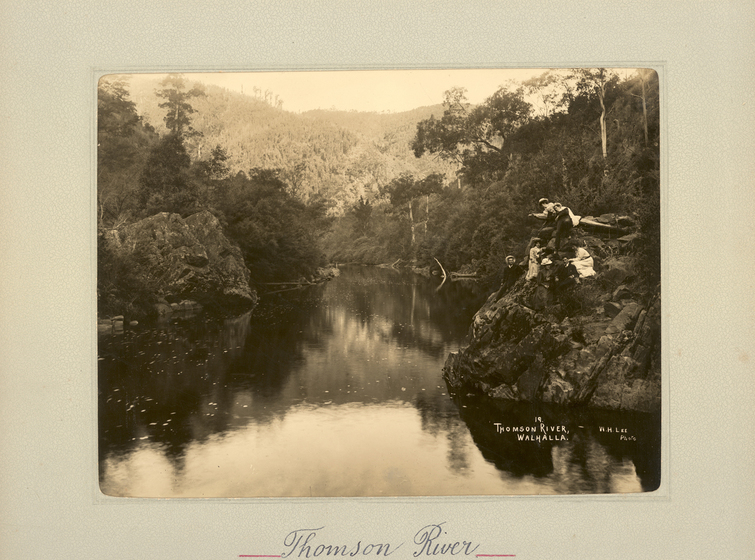 Black and white landscape photograph of a river, with tree-covered hills surrounding. On the right rocky bank sits a group of men and women. Text on the surrounding photo mount reads 'Thompson River'