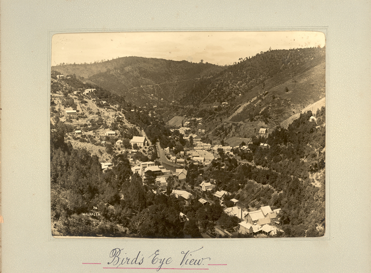 Black and white landscape photograph of a township sitting in a shallow gully, surrounded by tree-covered hills. . Text on the surrounding photo mount reads 'Birds Eye View'