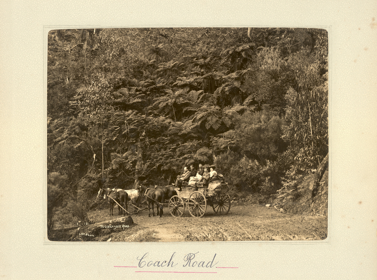 Black and white landscape photograph a cart drawn by four horses, with five people seated in it. The cart and horses are standing on a dirt road, with a tree-lined hillside behind. Text on the surrounding photo mount reads 'Coach Road'"