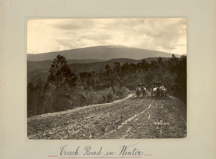 Dirt road with two horse-drawn carts carrying people in the distance. On the horizon are tree-covered hills. Text on the surrounding mount board reads, 'Coach Road in Winter'.