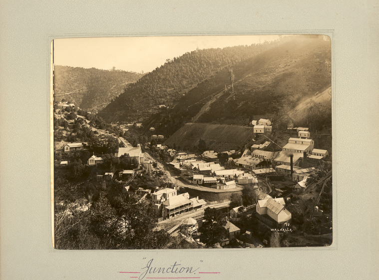 Black and white landscape photograph of a township set in a shallow gully, with tree-covered hills either side. Some buildings are placed up the hillside. Text on the surrounding photo mount reads 'Junction'