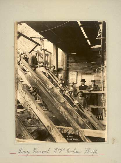Black and white portrait photograph of a timber room with one wall open, and lengths of timber beams on an angle towards the floor. Behind the beams stand a group of men. Text on the surrounding photo mount reads 'Long Tunnel Coy's Incline Shafts'