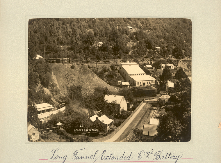 Black and white landscape photograph of the side of a tree-covered hill, with buildings set up the hill. A road is in the foreground.  Text on the surrounding photo mount reads 'Long Tunnel Extended Coy's Battery''