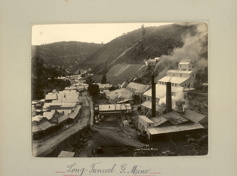 Black and white landscape photograph of white factory buildings and two smoke stacks, set at the base of and into the side of a hill. A road passes at the base. Text on the surrounding mount board reads 'Long Tunnel G. Mine;