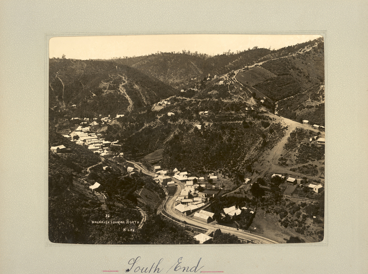 Black and white landscape photograph of a winding road running through a township at the bottom of a shallow gully. Sparsely tree-covered hills surround. Text on the surrounding mount board reads, 'South End'. 